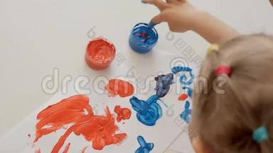 一个小可爱的女孩坐在桌子旁，用明亮的手指颜料画纸，把她的手指浸在颜料罐里。