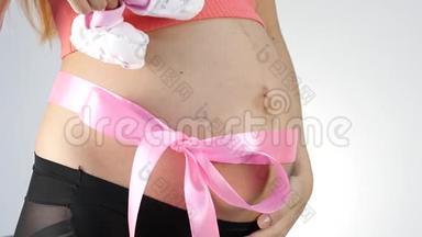 穿着内衣的怀<strong>孕妈妈</strong>在玩婴儿鞋。 腹部有粉红色的丝带。 慢动作