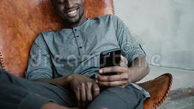 非洲男子坐在椅子上的肖像，使用智能手机。 人读<strong>留言</strong>，笑.. 打字按摩，微笑着