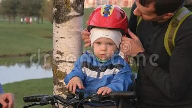 家长在公园里给幼儿戴上自行车头盔.. 一个男孩坐在自行车前的儿童座位上