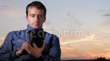 一个年轻人坐在日落时分，检查留言以检查平板电脑上的留言。 后面是美丽的天空。