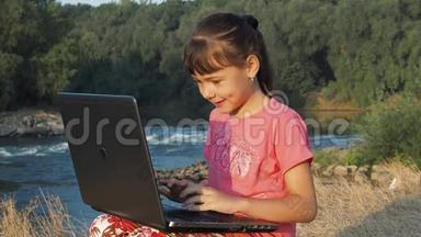 在河边拿着笔记本电脑的女孩。 河岸上的女孩带着笔记本电脑。 一个拿着笔记本电脑的小女孩