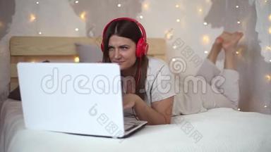 穿着睡衣的漂亮女孩<strong>用</strong>笔记本<strong>电脑</strong>在耳机上听音乐，心情很好地躺在床上。 高清高清
