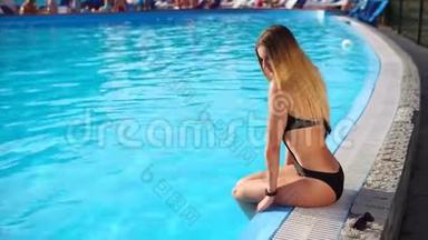 年轻苗条的金发女郎坐在游泳池附近，把她的腿放在水<strong>里</strong>。