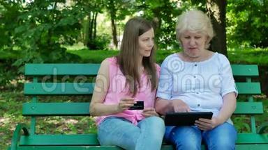 年<strong>长</strong>的女人和年轻的女人在公园里坐在<strong>长</strong>凳上网上买<strong>东西</strong>。 成年母亲和女儿通过