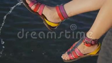 一个女孩的腿坐`水边。 那个女孩穿着彩色凉鞋在水边喋喋不休。
