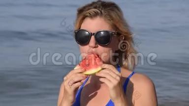 一个戴太阳镜的女人坐在海滩上吃西瓜。 一个成熟的西瓜在金发女郎手中