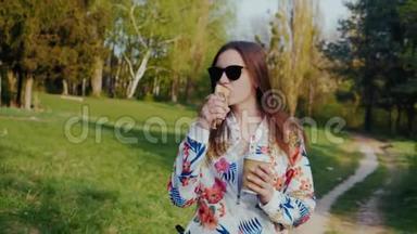 一个年轻的女人在公园里散步，<strong>吃</strong>一种食物，从纸杯里喝咖啡。 咖啡和<strong>热狗</strong>-去<strong>吃</strong>点心