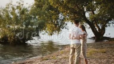 夕阳西下，一对年轻漂亮的夫妇站在河岸上。 迷人的男人和女人拥抱在水边。
