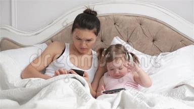 美丽的母亲和她可爱的女儿有两个马尾辫正在使用小玩意一起躺在白色的床上。 美丽美丽