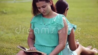 肖像。 一个漂亮的女孩和一个可爱的小女孩坐在草地上背靠背地看书，用平板电脑