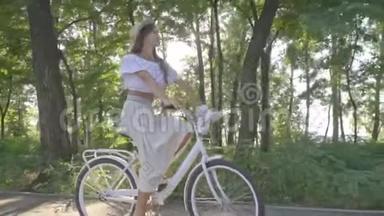 一件衬衫穿着一条白色裙子，漂亮的深色头发，沿着公园里的小路骑着一辆城市自行车，摘下她的草帽和草帽