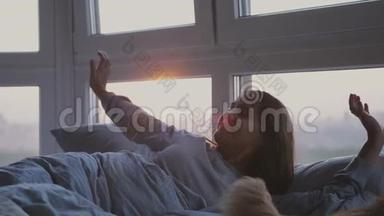 清晨，美丽的黑发女人醒来，躺在床上打哈欠，在日出时，在全景窗旁的房间里