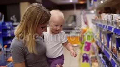 年轻的母亲戴着眼镜，抱着孩子，一边在超市的货架上挑选饼干