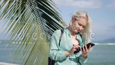 美丽的苗条女人，留着长长的金发和绿色衬衫，站在棕榈树附近，在<strong>后台</strong>使用智能<strong>手机</strong>