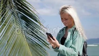 美丽的苗条女人，留着长长的金发和绿色衬衫，站在棕榈树附近，在<strong>后台</strong>使用智能<strong>手机</strong>