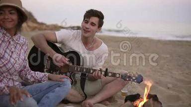 年轻人正和朋友一<strong>起坐</strong>在海滩上的火炉旁弹吉他。 他的女朋友带了烤