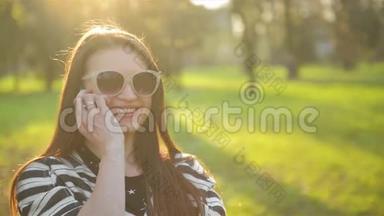 在阳光明媚的春天，带着长发和时尚太阳镜的浅黑色女孩正在公园里用手机聊天
