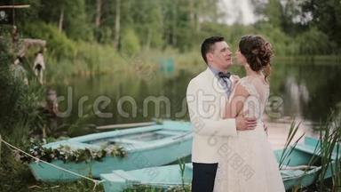 美丽的恋人在一个美丽的地方亲吻一个池塘，池塘上有蓝色的船。 新娘和新郎享受他们的婚礼