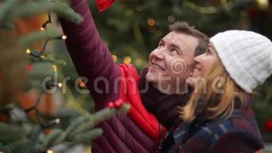 快乐夫妻在圣诞集市上选择圣诞树。 年轻的家庭在圣诞集市上聊天和接吻。 祝你快乐