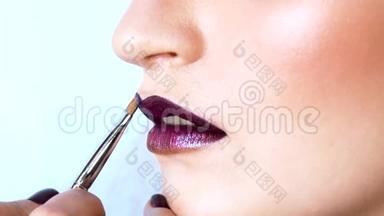 化妆师正在用刷子在模特的嘴唇上涂上明亮的紫色的小锡。 她的嘴微微张开.. 快关门。