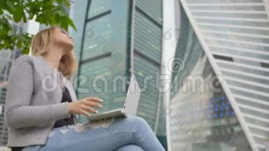 一个金发女孩在市中心和高楼的背景下使用白色笔记本电脑。 戴太阳镜的女孩。