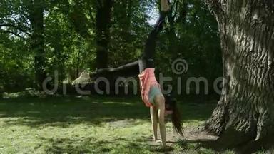 美丽的年轻女子在阳光公园练习瑜伽，倒立在树旁边。