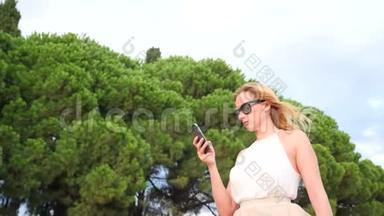 戴着太阳镜的优雅的金发女人对着天空和树叶打电话。 从下面看。 4k，慢