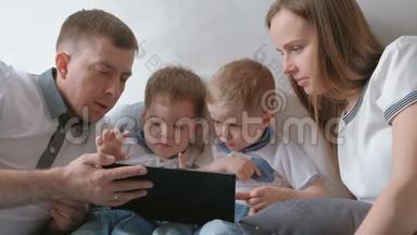 带平板<strong>电脑</strong>的家人。 妈妈，爸爸和<strong>两个</strong>儿子，双胞胎，蹒跚学步的孩子，看着躺在床上的平板<strong>电脑</strong>。