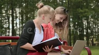 两个年轻漂亮的女学生手<strong>拿笔</strong>记本电脑在绿色公园的长凳上。 学习。 讨论。 侧视。