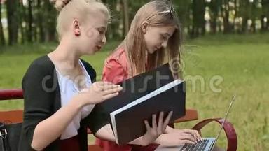 两个年轻漂亮的女<strong>学生</strong>手拿笔记本电脑在绿色公园的长凳上。 学习。 <strong>讨论</strong>。 侧视。