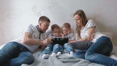 带平板电脑的家人。 妈妈，爸爸和两个儿子，双胞胎，蹒跚学步的孩子，看着躺在床上的平板电脑。