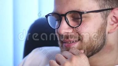 一个戴眼镜的大胡子年轻人坐在<strong>办公室用电脑</strong>工作的画像。 <strong>电脑</strong>屏幕显示