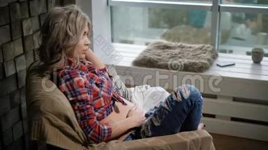 年轻漂亮的孕妇穿着格子衬衫和蓝色牛仔裤坐在靠近窗户的扶手椅上。 她在吹她的头发
