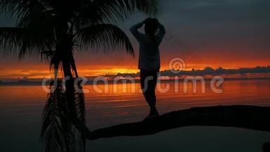 一个女孩站在海滩上红日落的背景下的棕榈树上