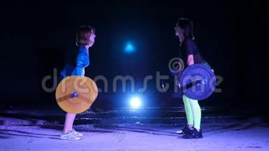 两个运动女孩，运动员，举起<strong>杠铃</strong>，用<strong>杠铃</strong>做仰卧起坐.. 晚上，<strong>在</strong>探照灯的照射下，