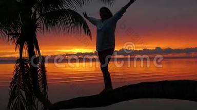 一个女孩站在海滩上红日落的背景下的棕榈树上