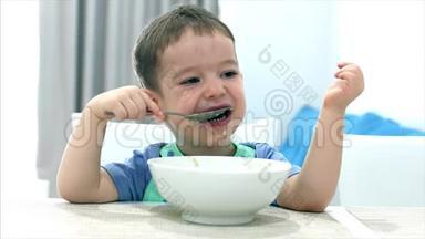 小可爱的孩子坐在一张桌子旁，吃自己的燕麦片，孩子吃得很自愿。 快乐童年的概念。