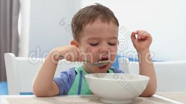 小可爱的孩子坐在一张桌子旁，吃自己的燕麦片，孩子吃得很自愿。 快乐童年的概念。