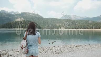 年轻女子用智能手机在山湖前拍照。 美丽的白种人女孩在一个女孩子中度过的时光