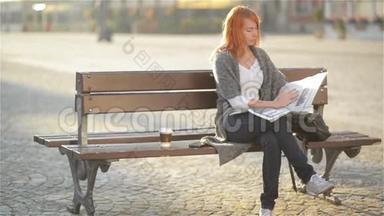 微笑的女人，红头发，坐在长凳上，喝咖啡，看报纸，阳光明媚的一天