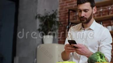 男人在家厨房用手机浏览。 英俊的年轻人在智能手机上浏览，微笑着快乐。 一个男人吃