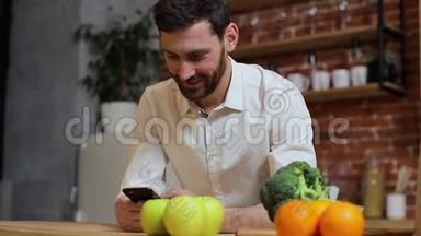 男人在家厨房用手机浏览。 英俊的年轻人在智能手机上浏览，微笑着快乐。 一个男人吃
