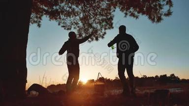 两个男人在一个野营聚会上跳舞，在大自然上，有趣的阳光剪影。 两个烧烤的游客休息一下