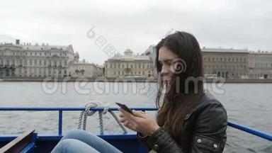 美丽的黑发女孩使用她的智能手机坐在河边公共汽车。 水，背景建筑，慢莫