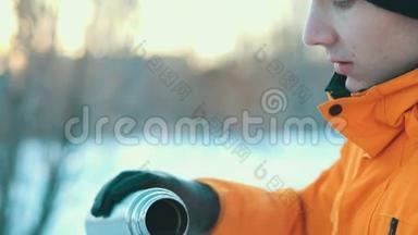 一个年轻人在冬天的公园里<strong>喝热水</strong>瓶里的热饮料。 大雪纷飞。 穿着橙色夹克和温暖衣服的人