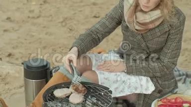 穿着外套的年轻女子坐在河边的海滩上，大海，野餐，在烤架上煮肉，寒冷的天气