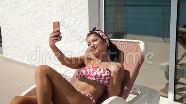 微笑的女人躺在躺椅上，在智能手机上自拍。