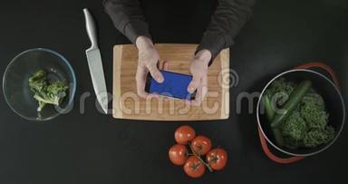 桌面上方手持手机的手的俯视图。厨房概念。砧板、刀和蔬菜