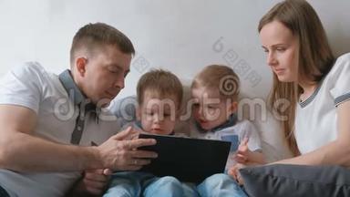 带平板电脑的家人。 妈妈，爸爸和两个儿子，双胞胎，蹒跚学步的孩子，看着躺在床上的平板电脑。